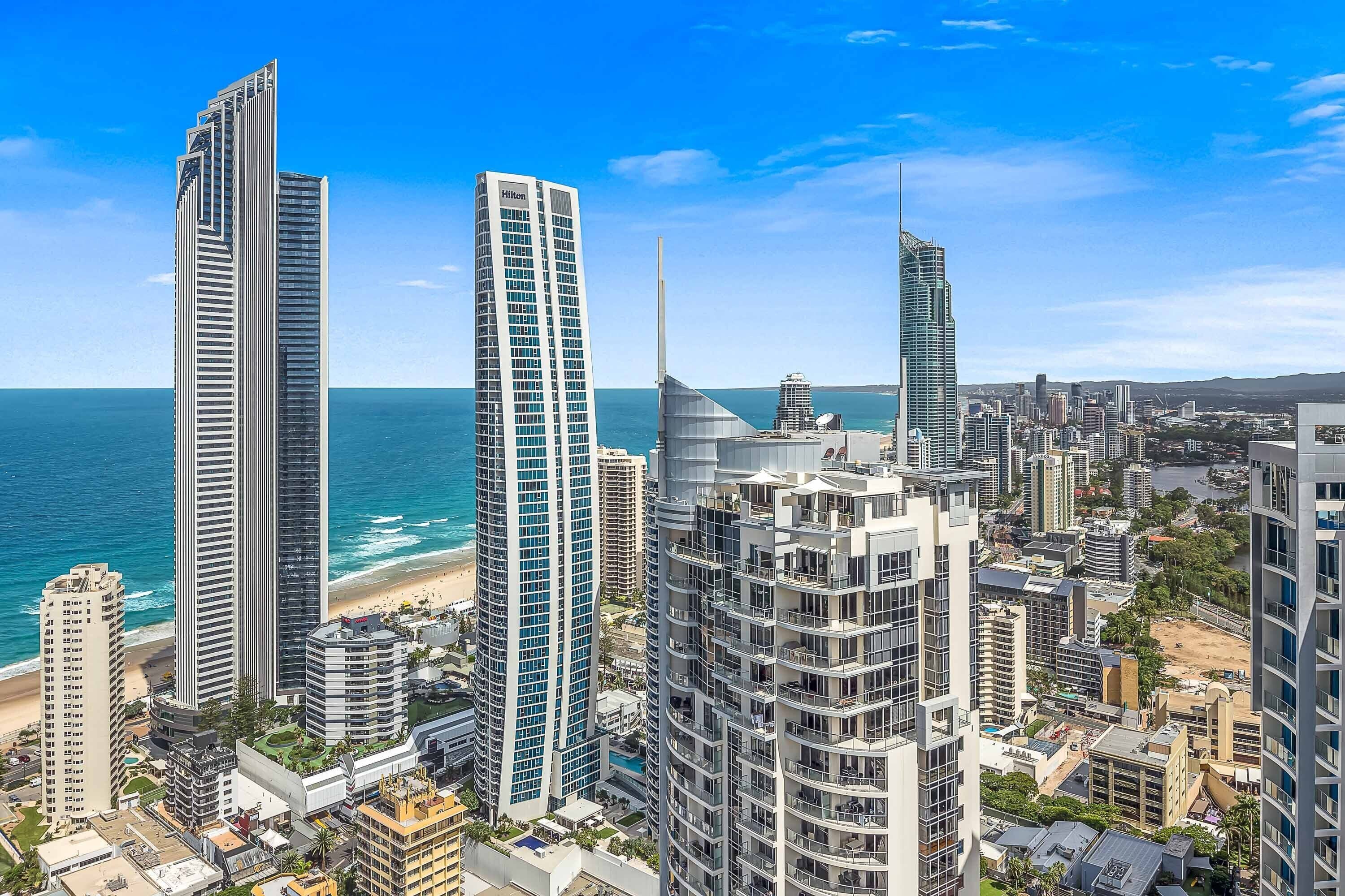 Un paisaje urbano costero con edificios altos, incluyendo un rascacielos destacado con un diseño único y una playa al fondo.
