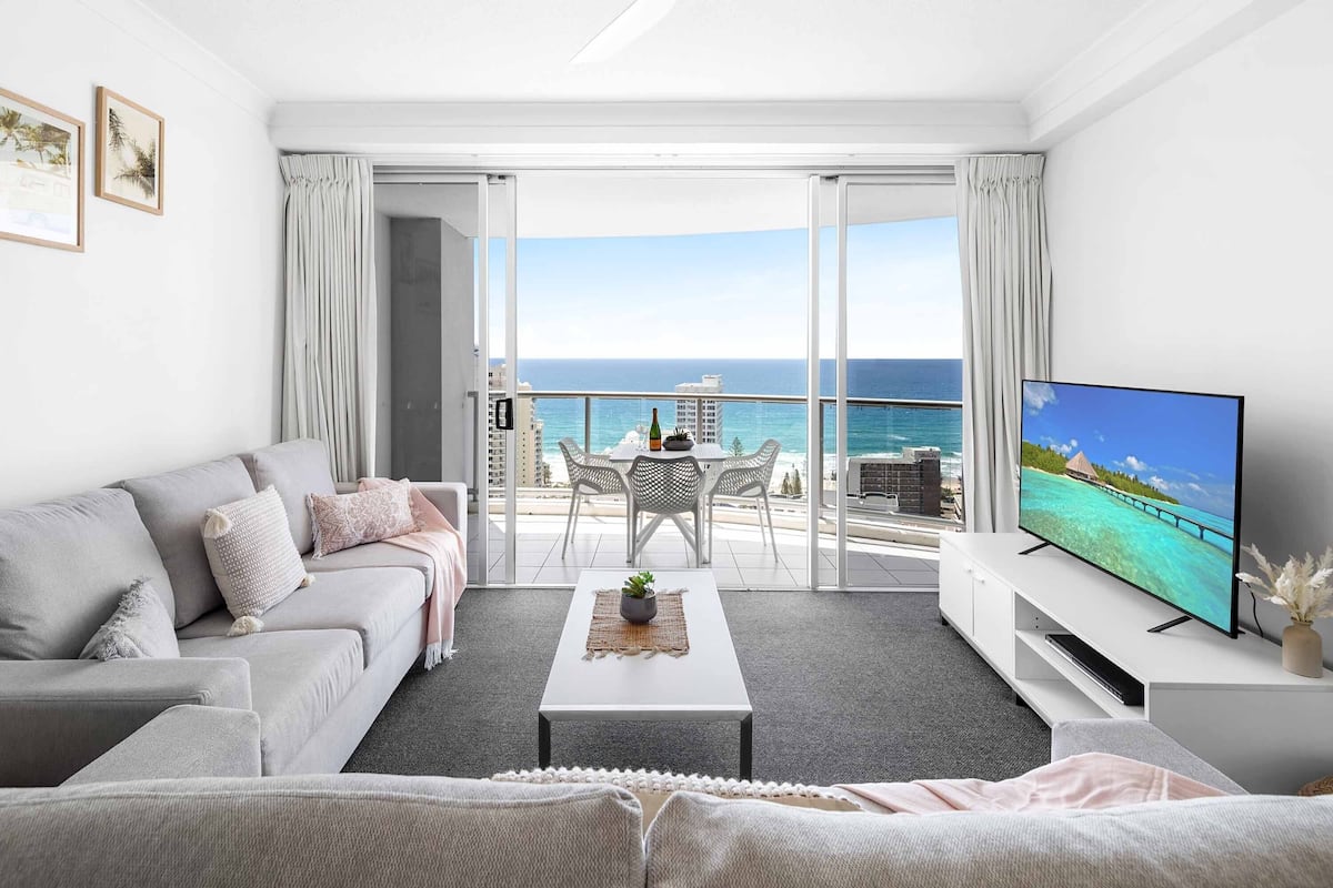 A modern living room with a sofa, coffee table, and TV, overlooking a balcony with a view of the ocean.