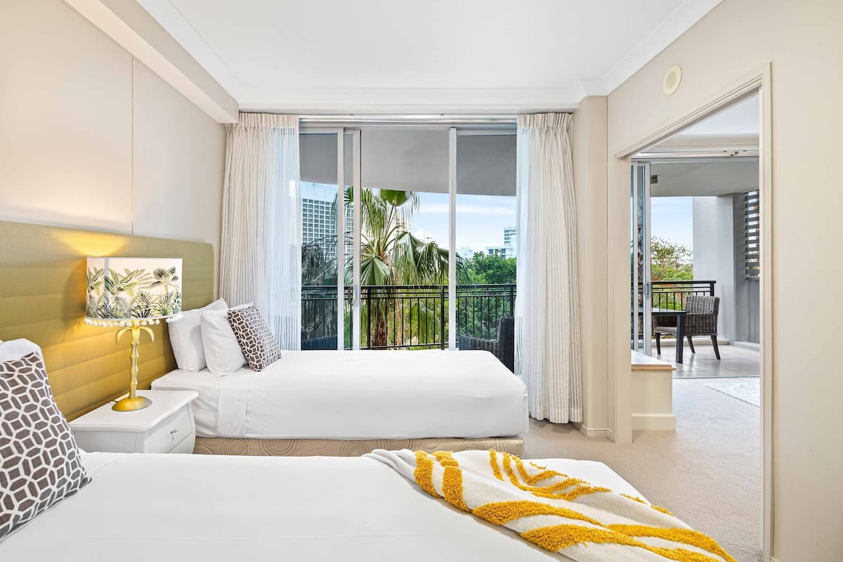A hotel room with two beds, a nightstand with a lamp, a view of a balcony with outdoor furniture, and a sliding glass door leading to an outdoor area.