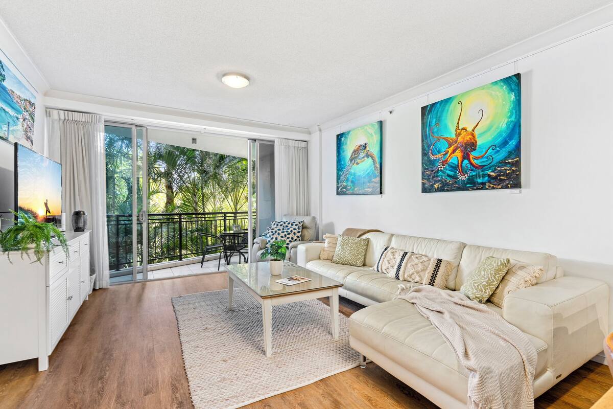 A modern living room with a glass coffee table, a beige sofa, and two large paintings.