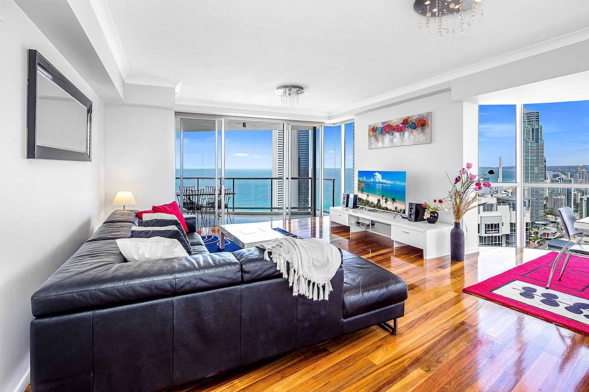 A modern living room with a black leather sofa, a white entertainment unit, and a large window overlooking the city.