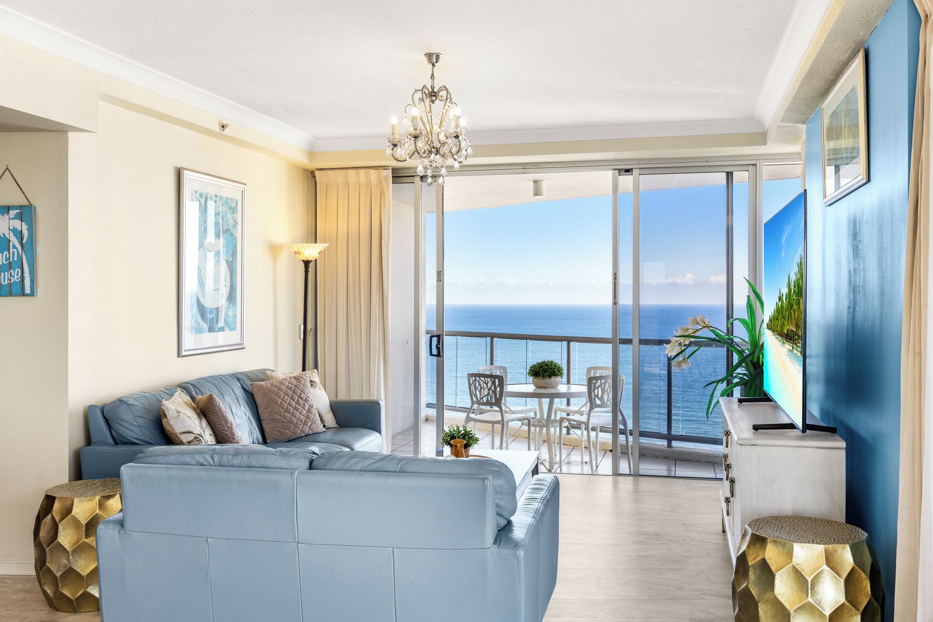 A modern living room with a sofa, a dining table, and chairs, a chandelier, and a large window with a view of the sea.