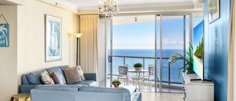 A modern living room with a sofa, a dining table, and chairs, a chandelier, and a large window with a view of the sea.