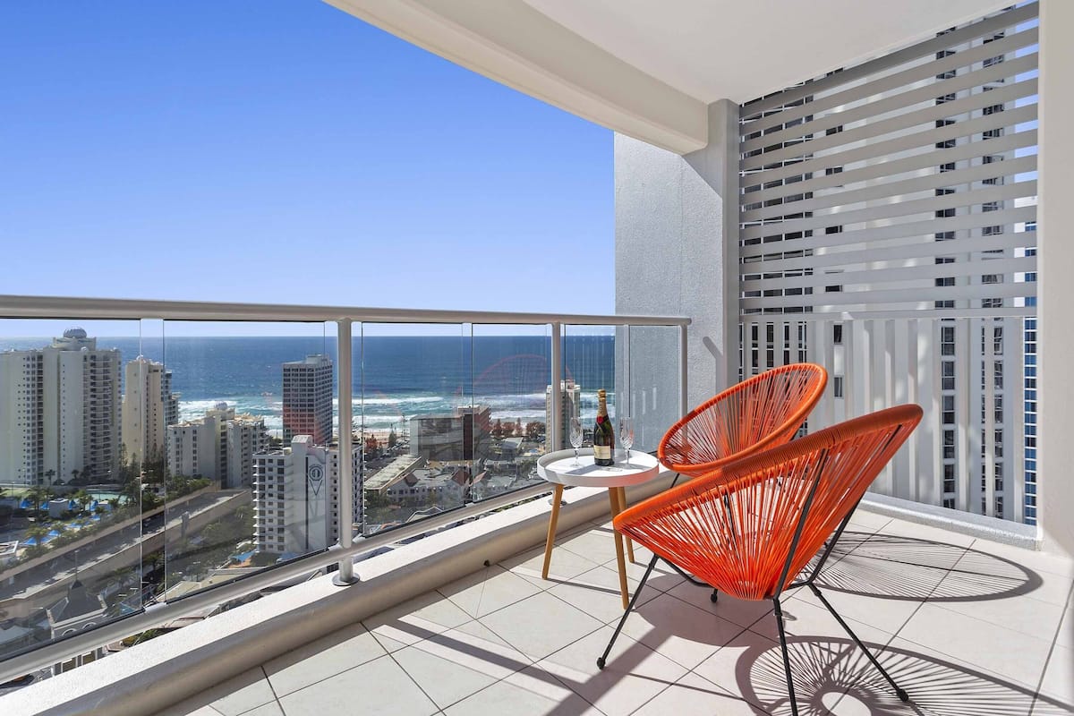 A balcony with a view of the ocean and cityscape, featuring two orange chairs and a small table with a bottle and glass.