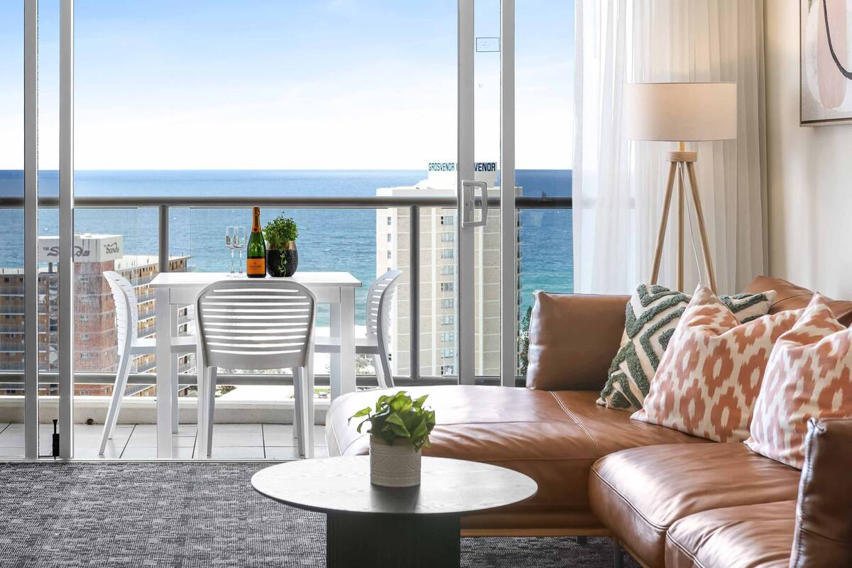 A balcony with a view of the sea, a brown leather sofa, a small table with a plant, and a white chair.