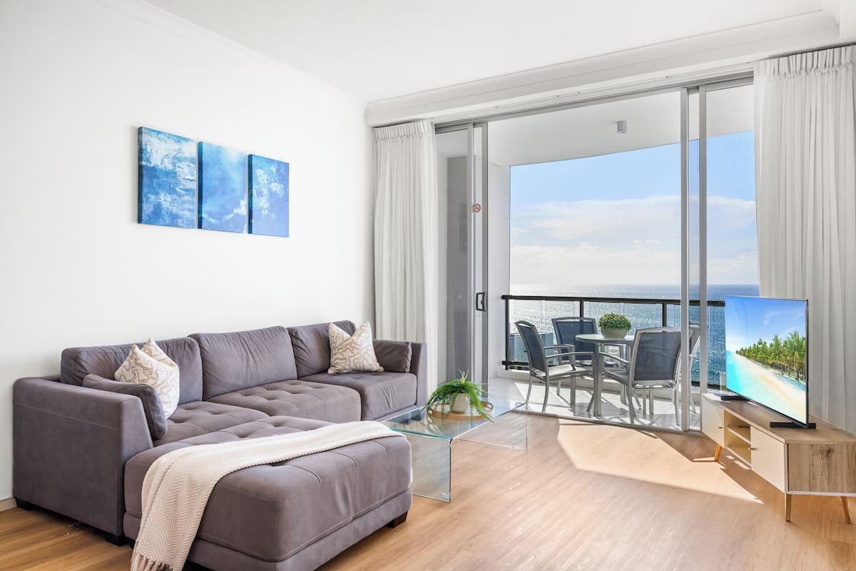 A modern living room with a grey sofa, a glass coffee table, and a balcony with a view of the sea.