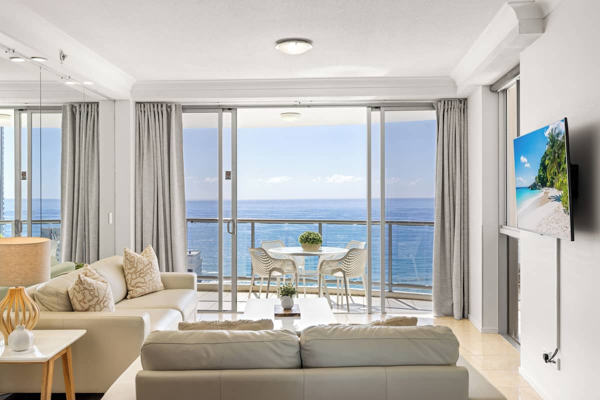 A modern living room with a balcony overlooking the sea, featuring a sofa, a table with chairs, and a television.