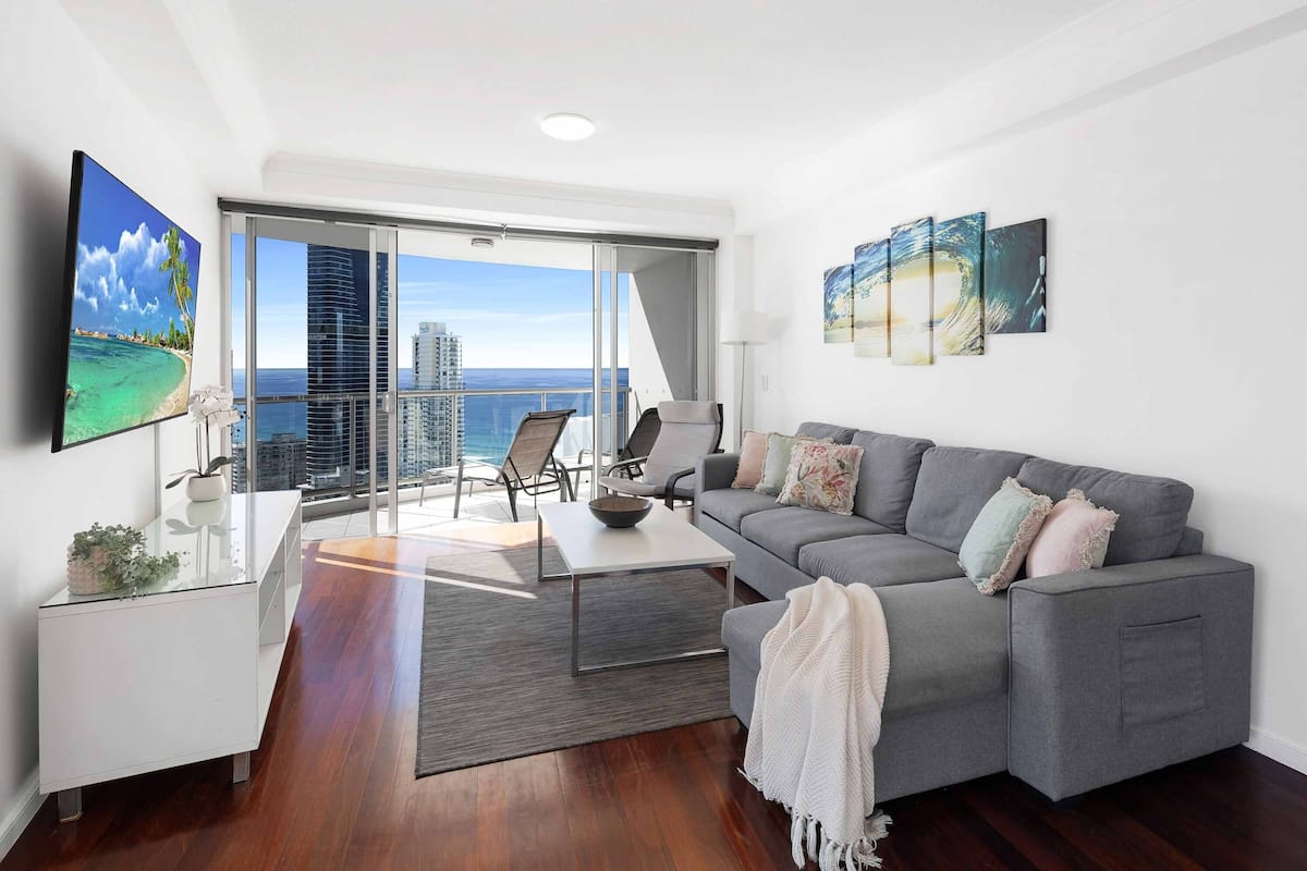 A modern living room with a grey sofa, a flat-screen TV, and a balcony with a view of the city and ocean.