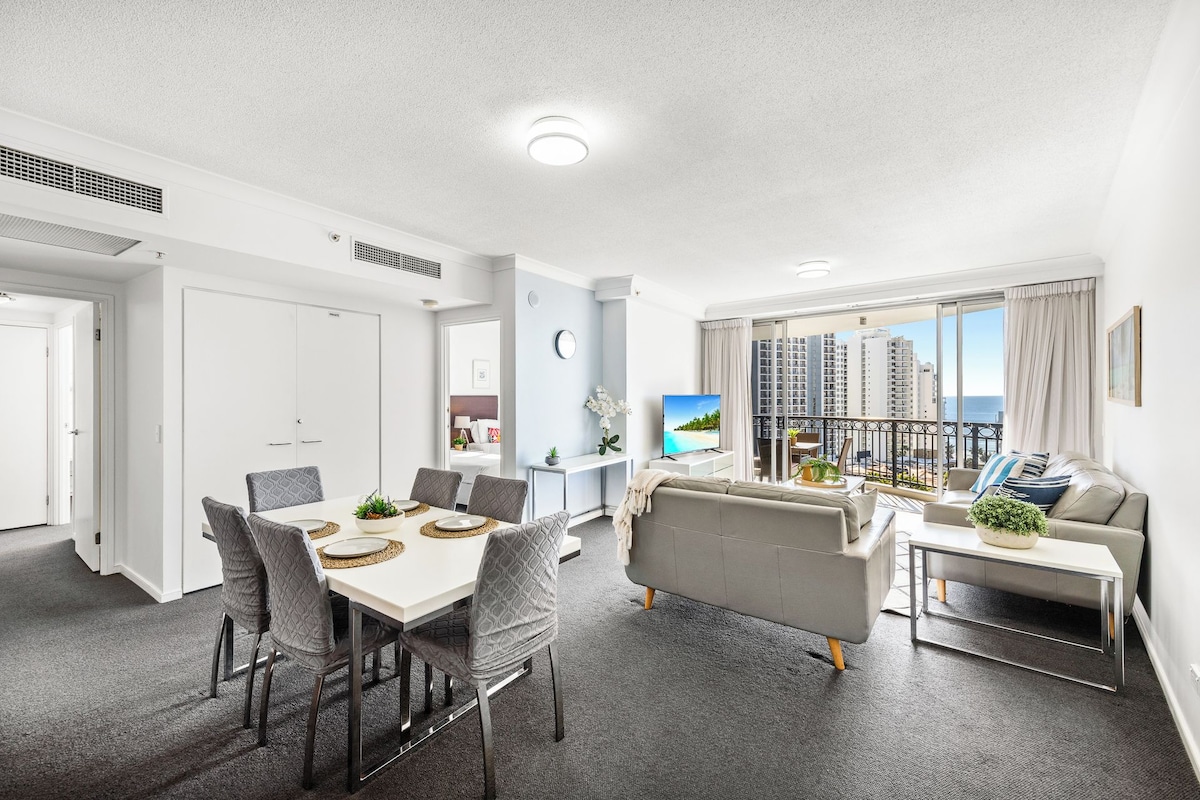 A modern living room with a grey sofa, a dining table with chairs, a TV, and a balcony with a city view.