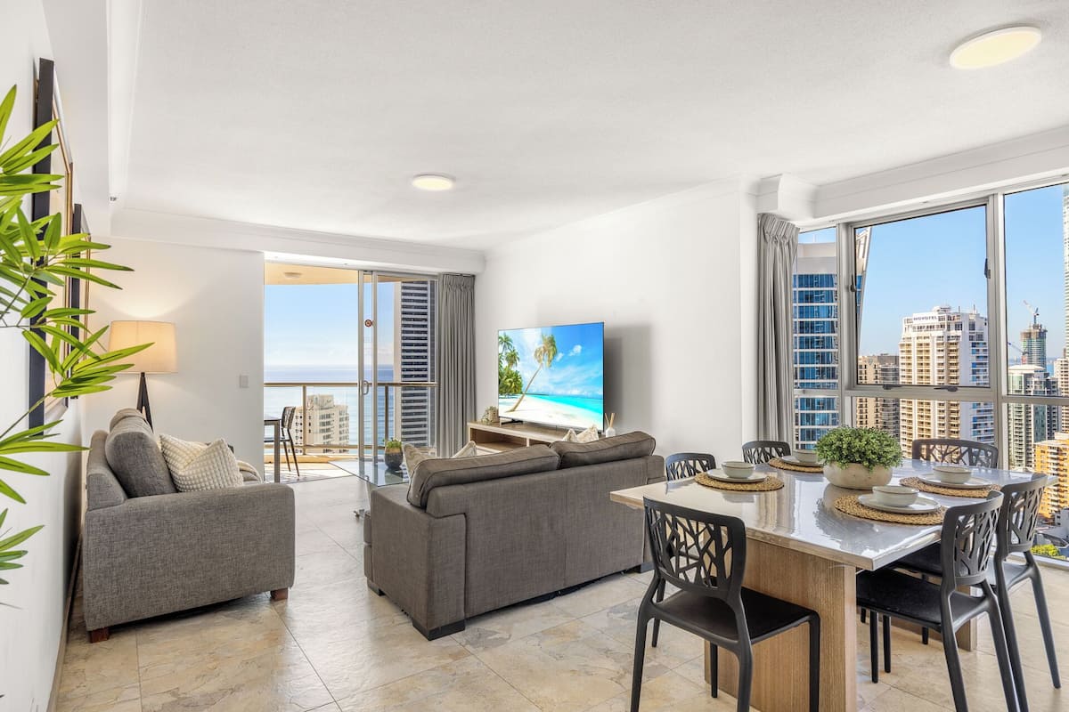 A spacious living area with a balcony, a flat-screen TV, and a dining table with chairs.
