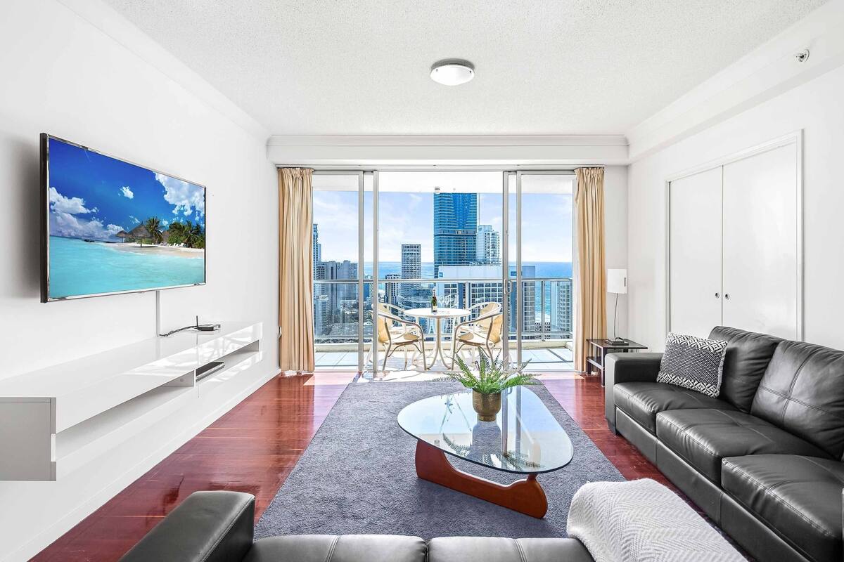 A modern living room with a large TV, a glass coffee table, a leather sofa, and a balcony with a city view.