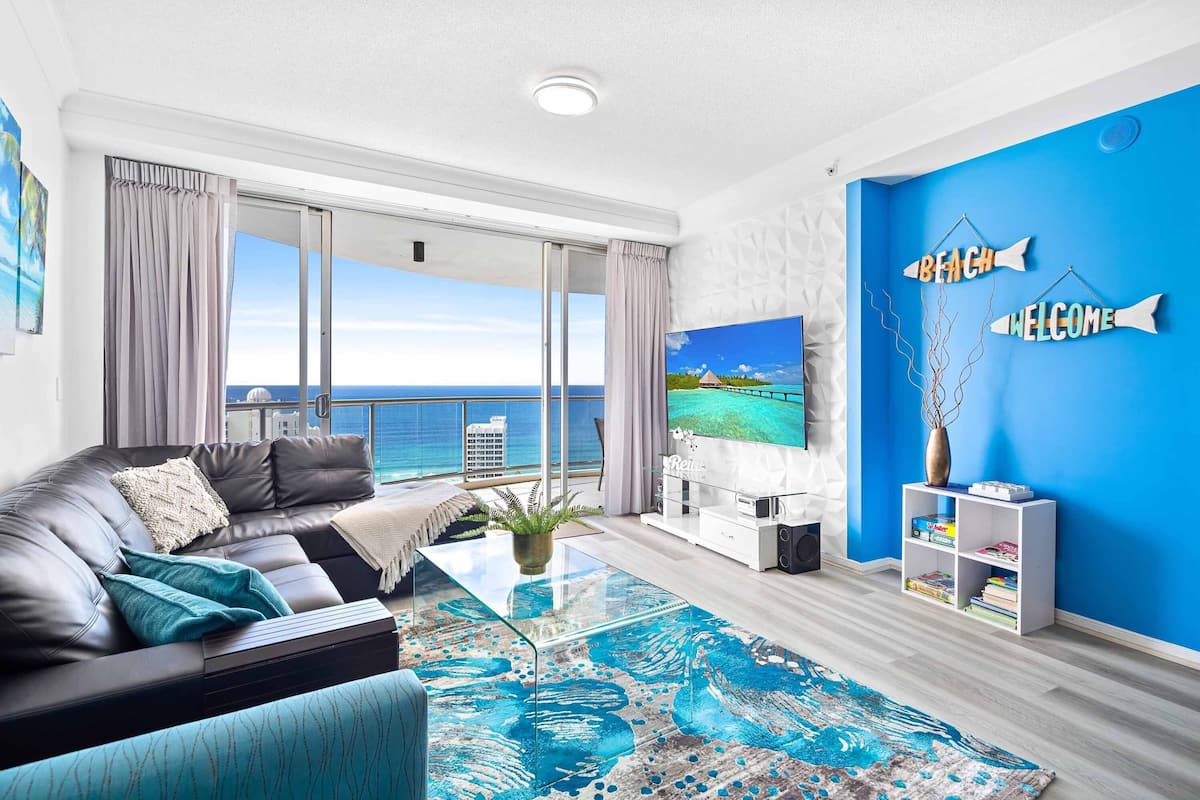 A modern living room with a large window overlooking the sea, a black leather sofa, a glass coffee table, and a blue accent wall with 'BEACH' and 'WELCOME' signs.