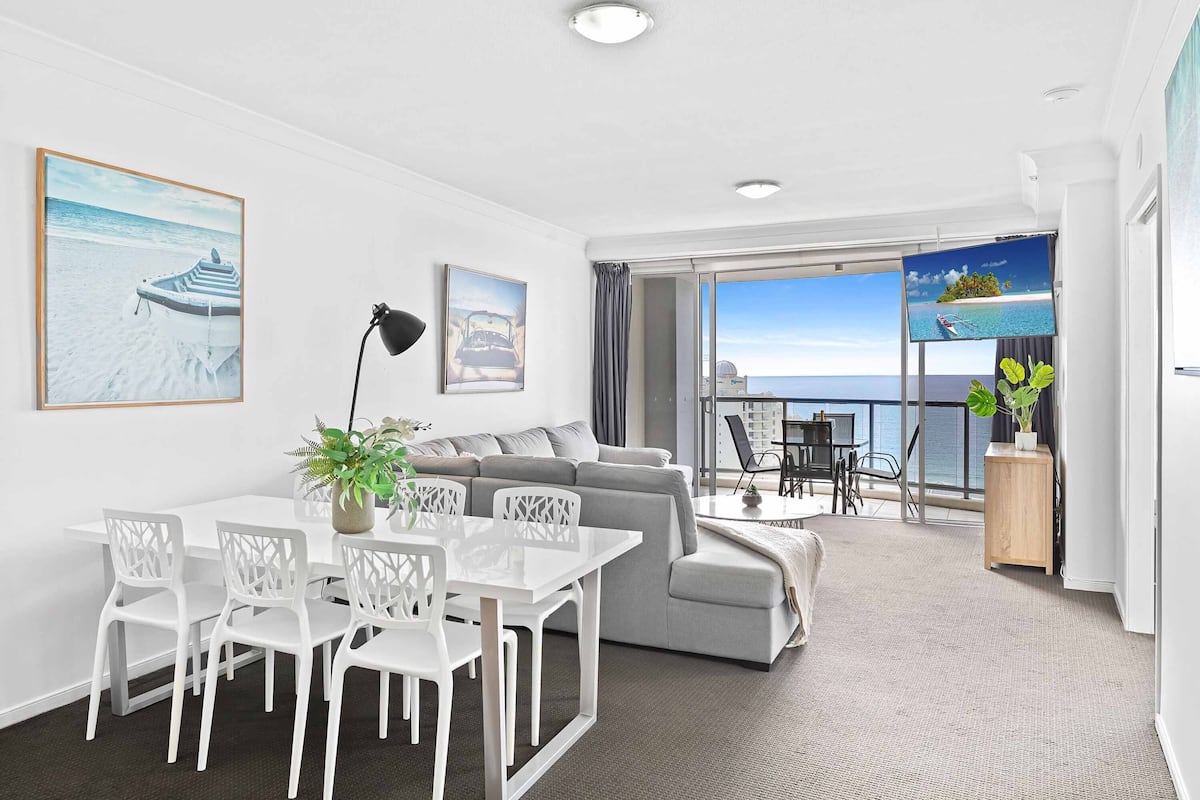 A modern living room with a dining area, a sofa, a TV, and a balcony with a view of the sea.