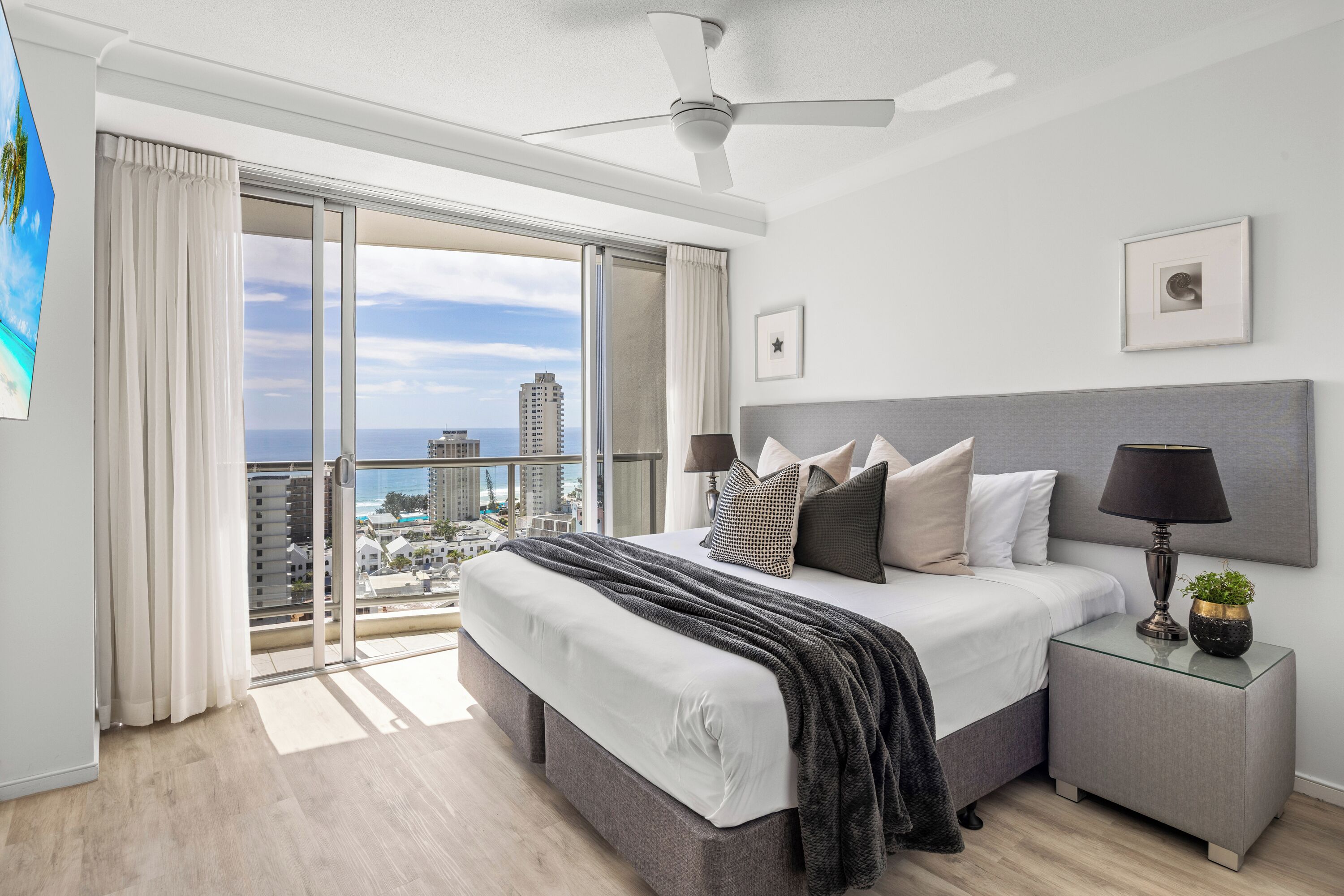 A modern bedroom with a large bed, a nightstand, a ceiling fan, and a view of the city through a sliding glass door.