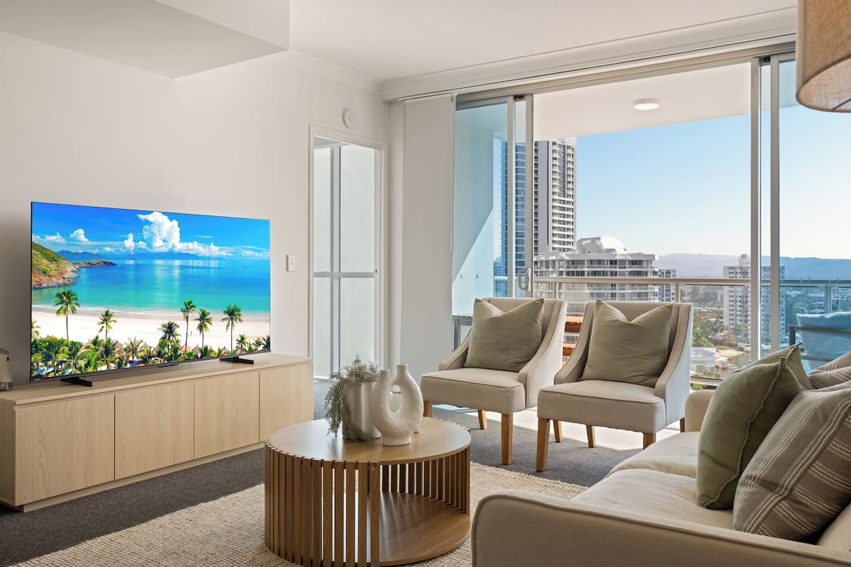 A modern living room with a large flat-screen TV, comfortable seating, and a view of a cityscape through large windows.