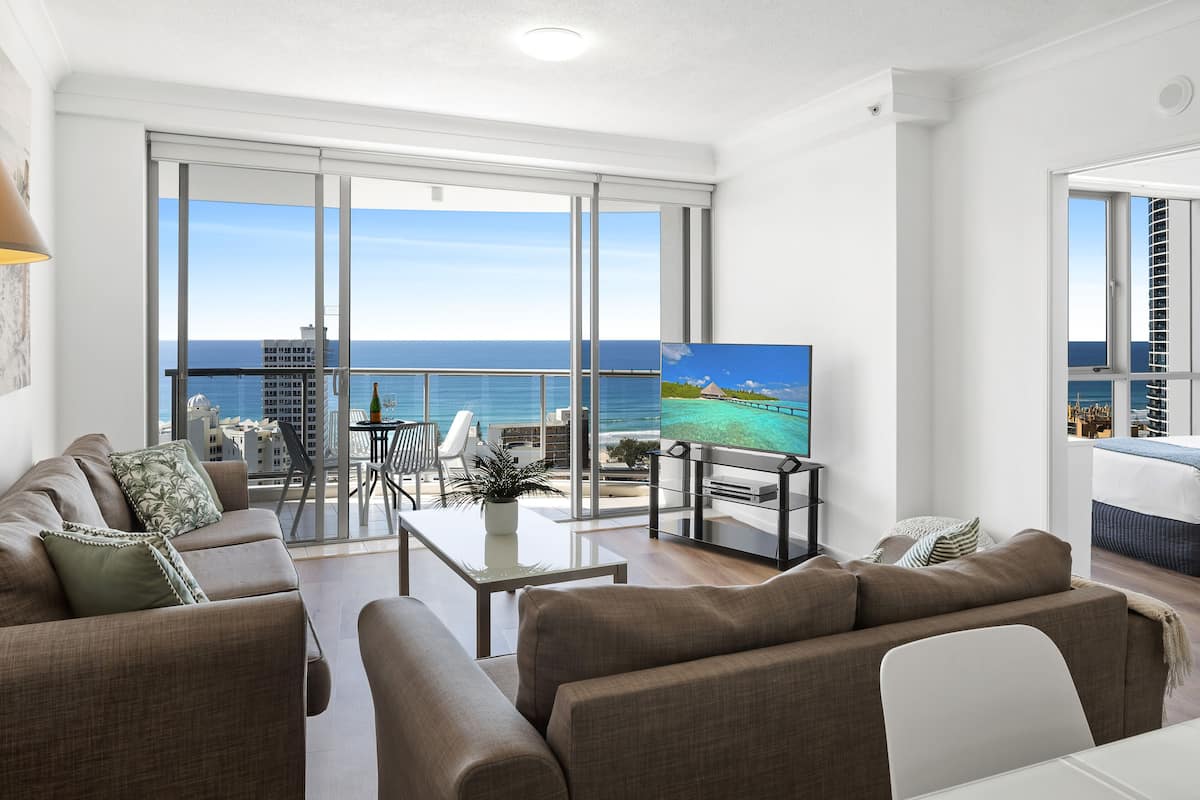 A modern living room with a large TV, comfortable seating, a balcony with a view of the sea, and a dining area.