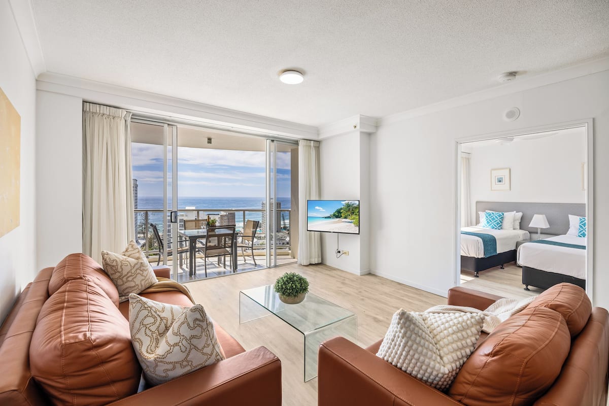 A modern hotel room with a balcony, a glass coffee table, leather sofas, and a bed with blue and white bedding.