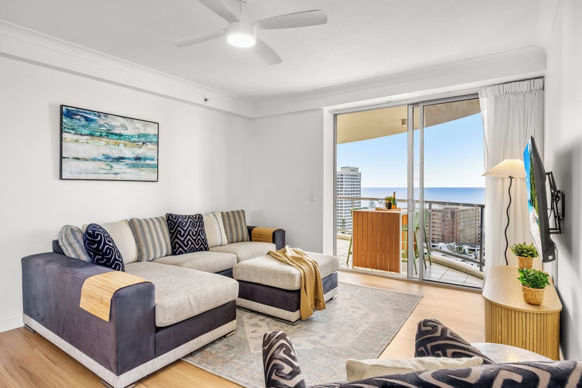 A modern living room with a sectional sofa, a coffee table, and a view of the cityscape through large sliding doors.