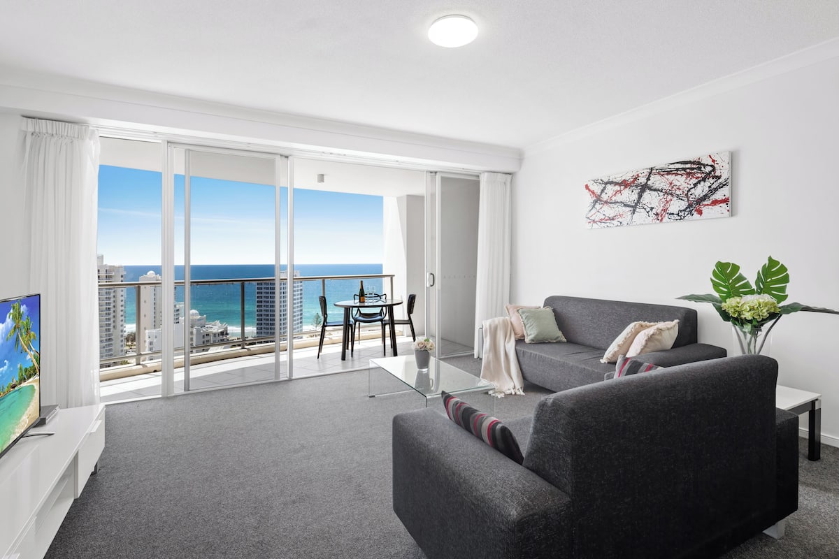 A modern living room with a grey sofa, a glass coffee table, and a view of the ocean.