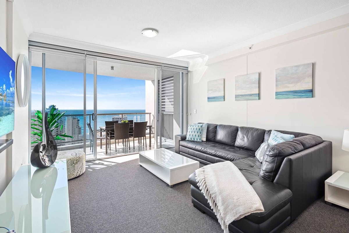 A modern living room with a black leather sofa, a glass coffee table, and a balcony with a view of the sea.