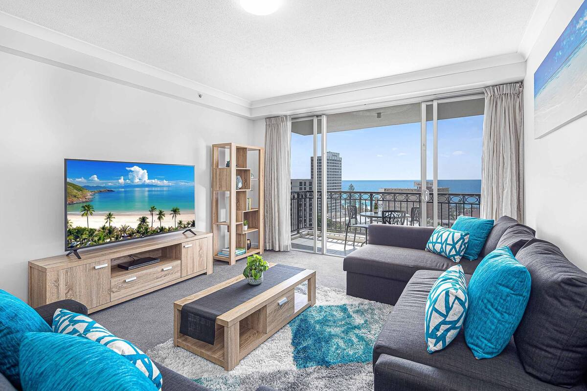 A modern living room with a large flat-screen TV, a wooden entertainment unit, a grey sofa with blue cushions, and a view of the ocean through large sliding doors.