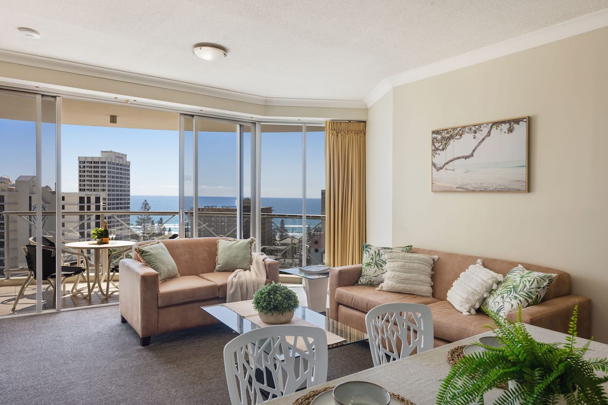 A modern living room with a large window overlooking a cityscape and the sea. There are two sofas, a dining table, and a small balcony area with a table and chairs.