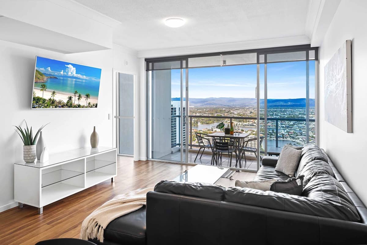 A modern living room with a large TV, a sofa, a dining table, and a balcony with a city view.