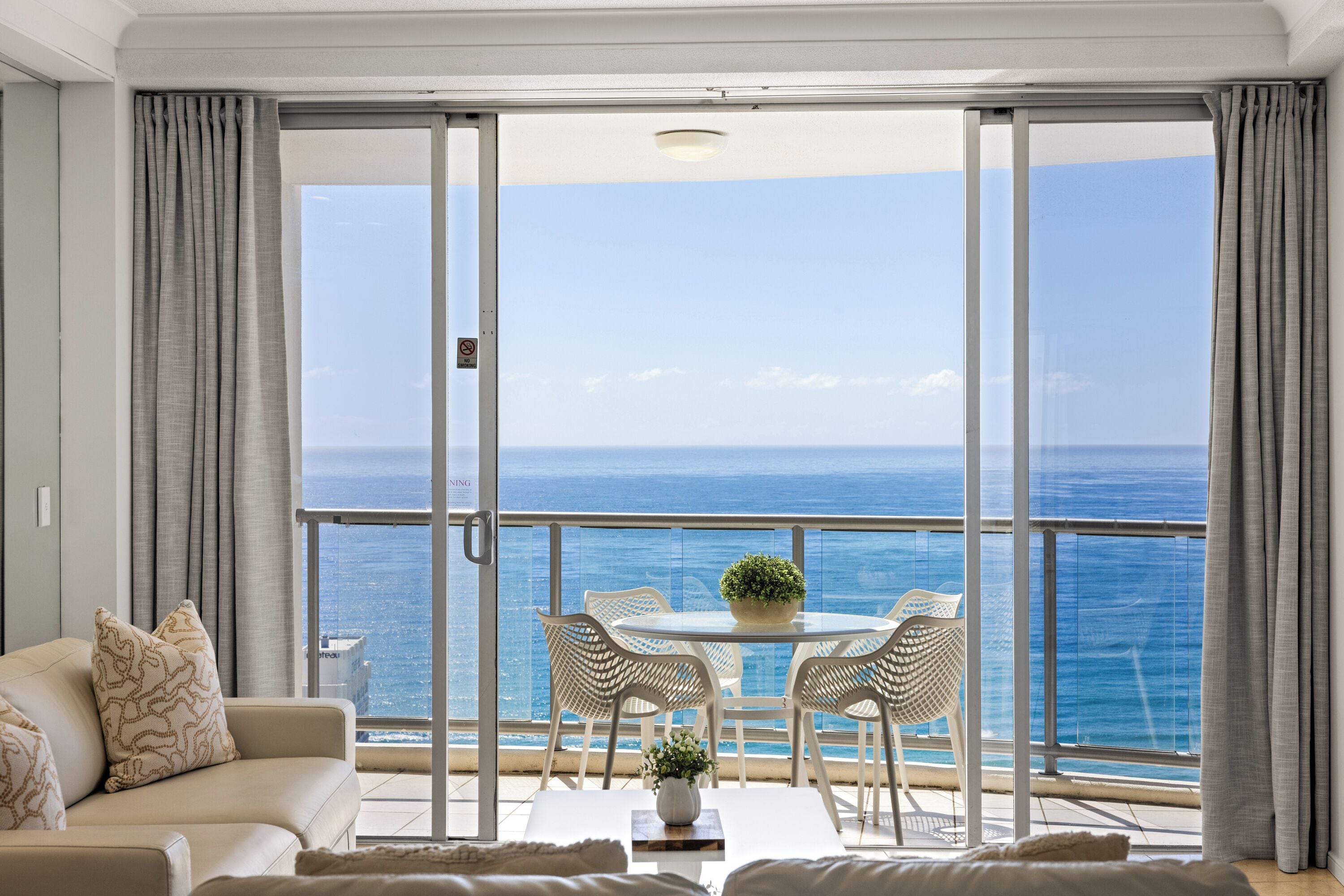 A balcony with a glass door, a white sofa, a round table with chairs, and a view of the ocean.