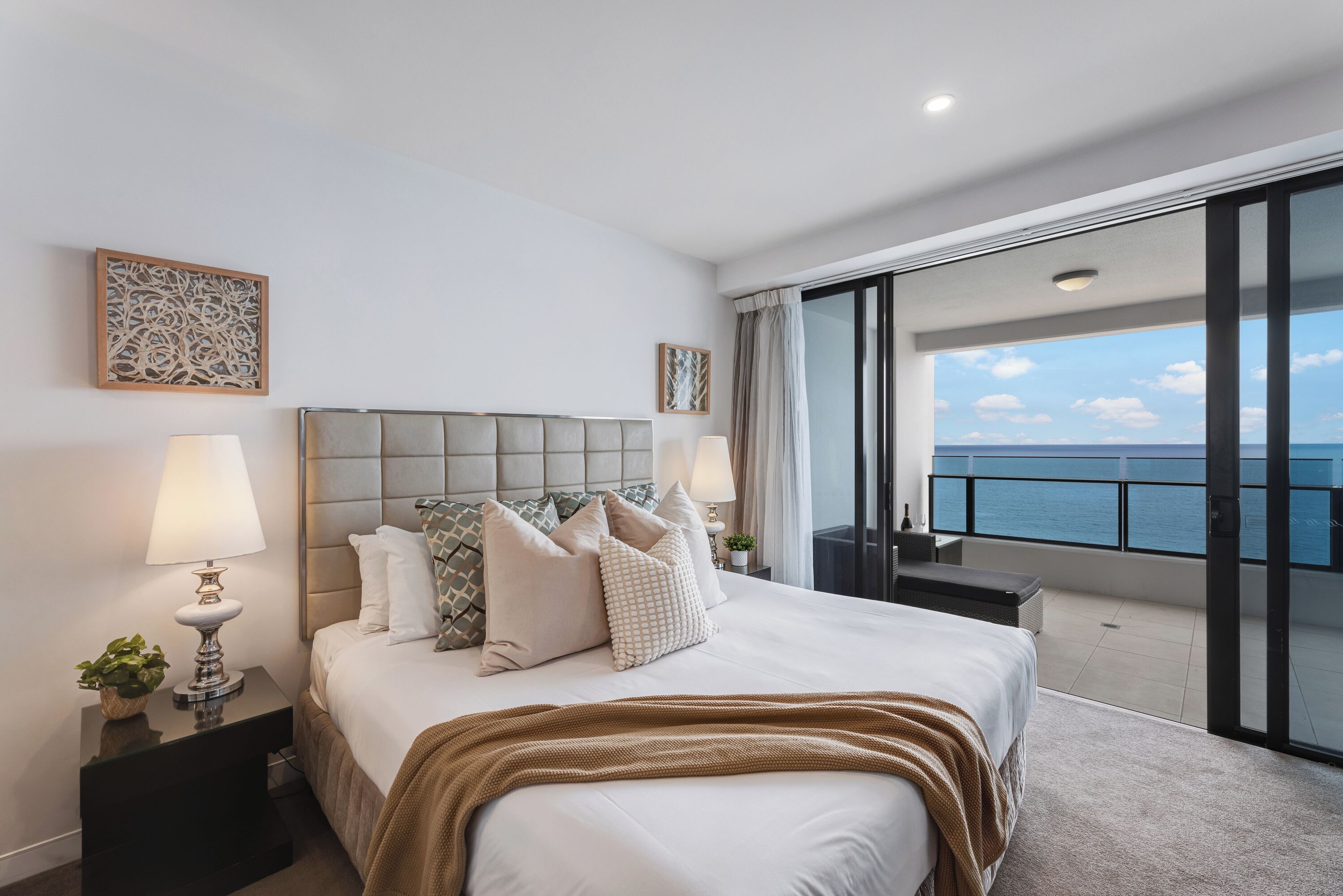 A hotel room with a large bed, two bedside tables with lamps, a view of the sea through a balcony, and a decorative wall piece.