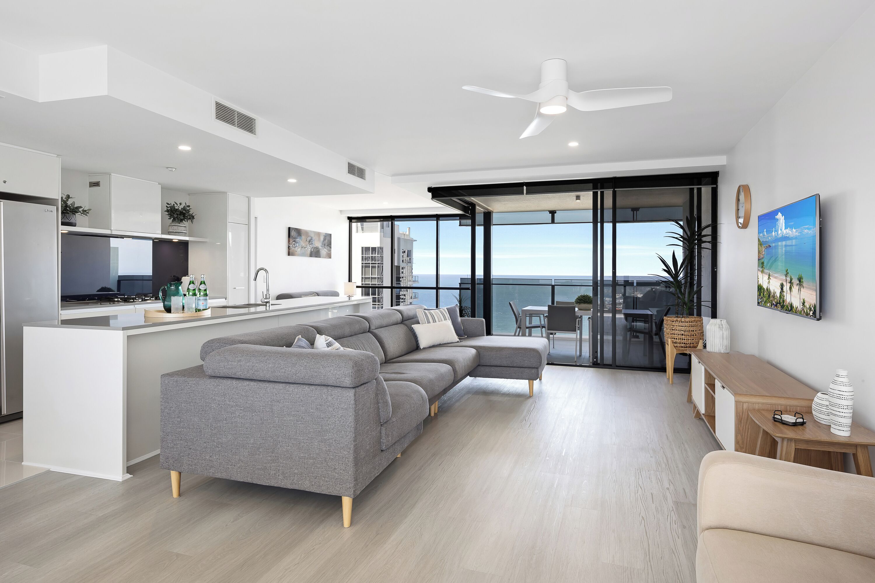 A modern living room with a grey sectional sofa, a wooden coffee table, and a balcony with a view of the sea.