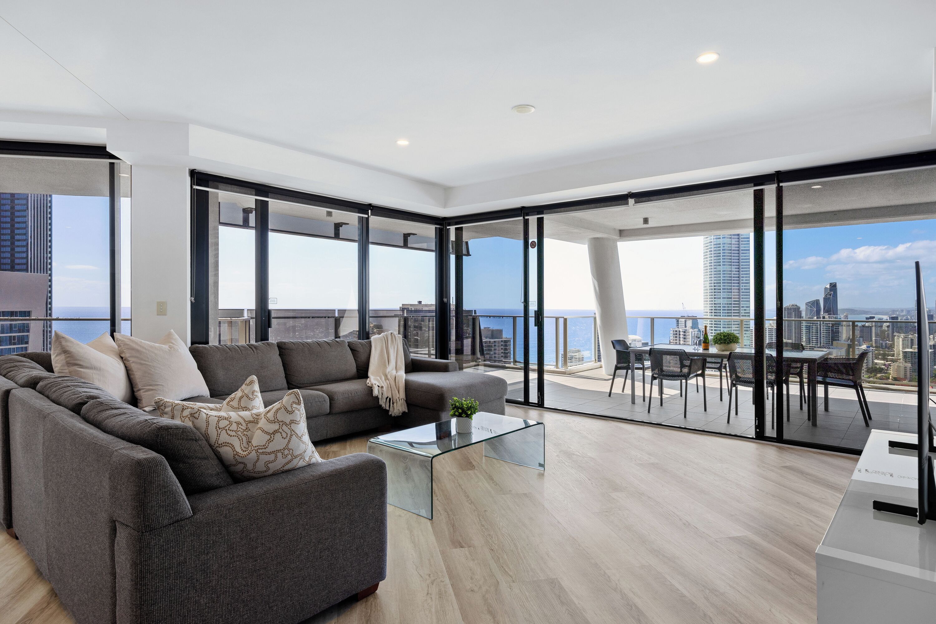 A modern living room with a grey sofa, a glass coffee table, and a city view through large windows.
