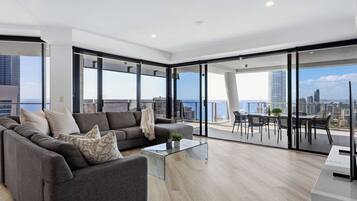 A modern living room with a grey sofa, a glass coffee table, and a city view through large windows.