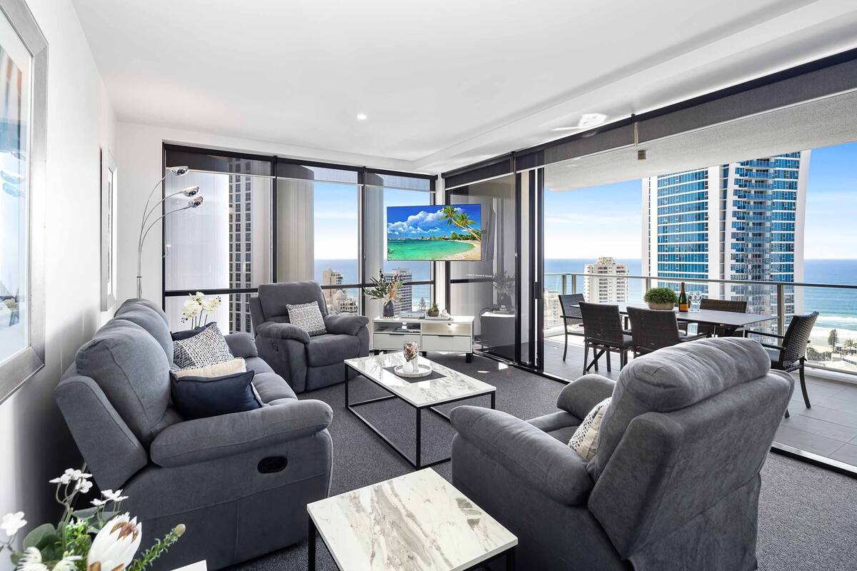 A modern living room with grey sofas, a coffee table, and a TV displaying a beach scene. Large windows offer a view of the ocean and high-rise buildings.