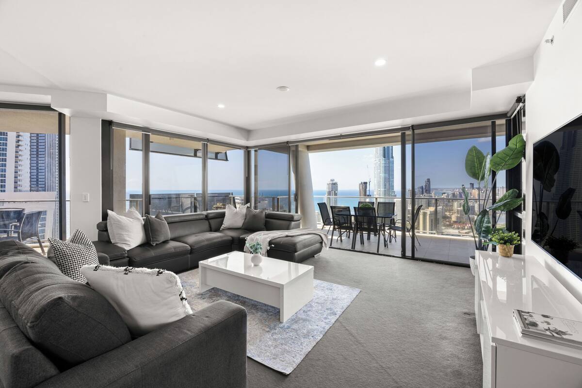A modern living room with a large sofa, a coffee table, and a TV. There is a balcony with a city view.