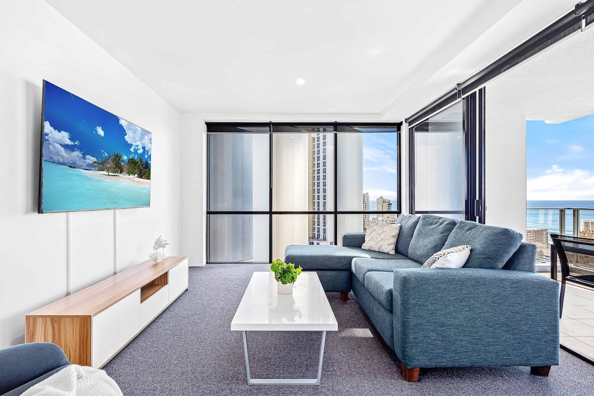 A modern living room with a grey sofa, a white coffee table, and a large window offering a view of the ocean and buildings.
