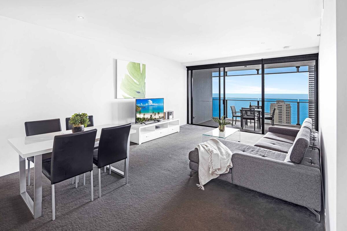 A modern living room with a white table, black chairs, a grey sofa, and a TV on a white stand. There is a large window with a view of the ocean and buildings.