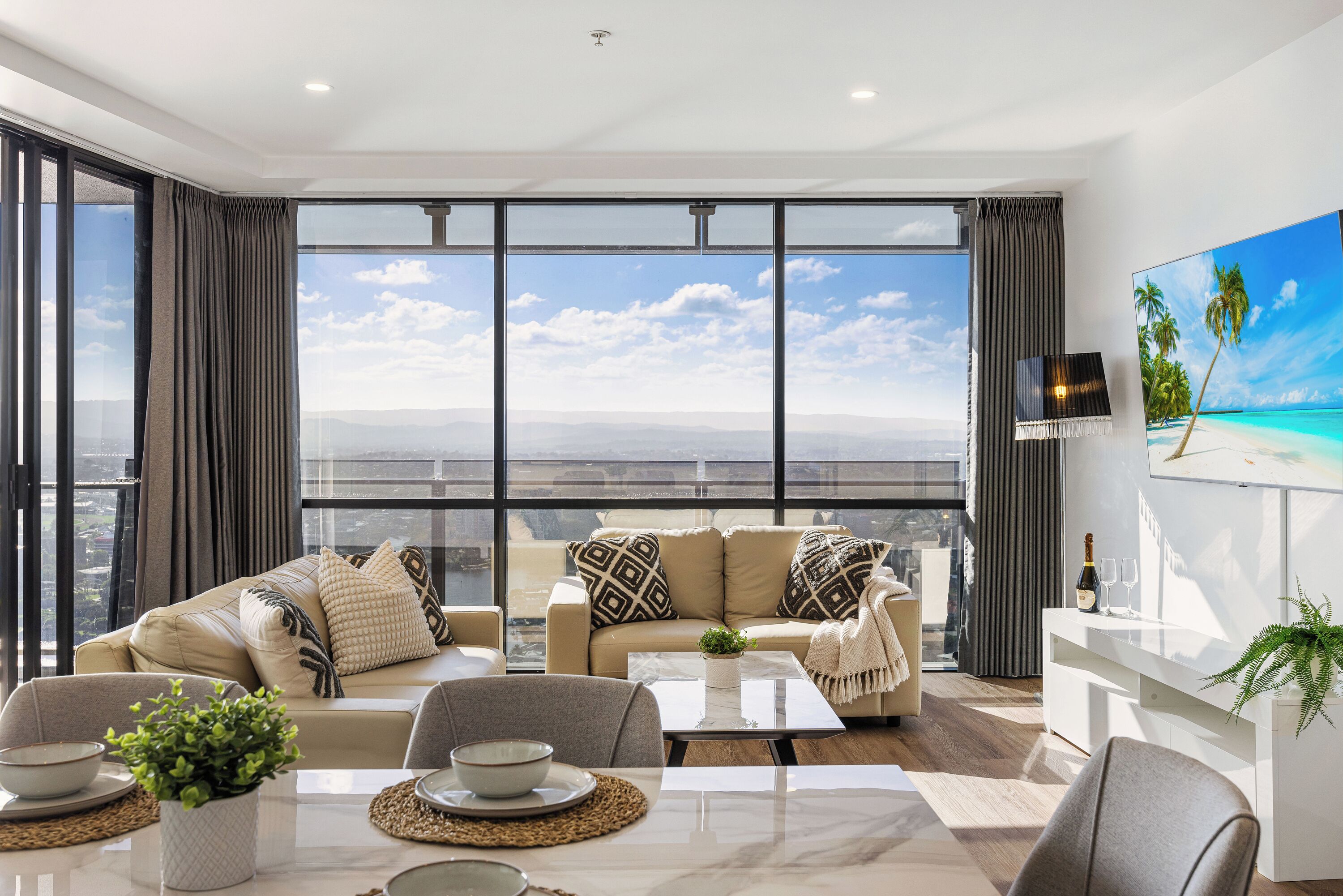 A modern living room with a large window, a sofa, a coffee table, and a TV on the wall.