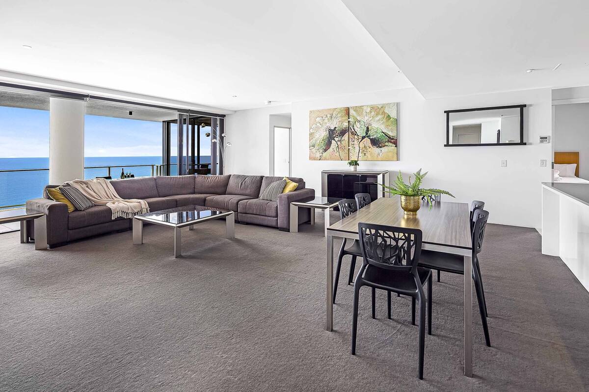 A modern living room with a large sofa, a dining table, and a view of the sea.