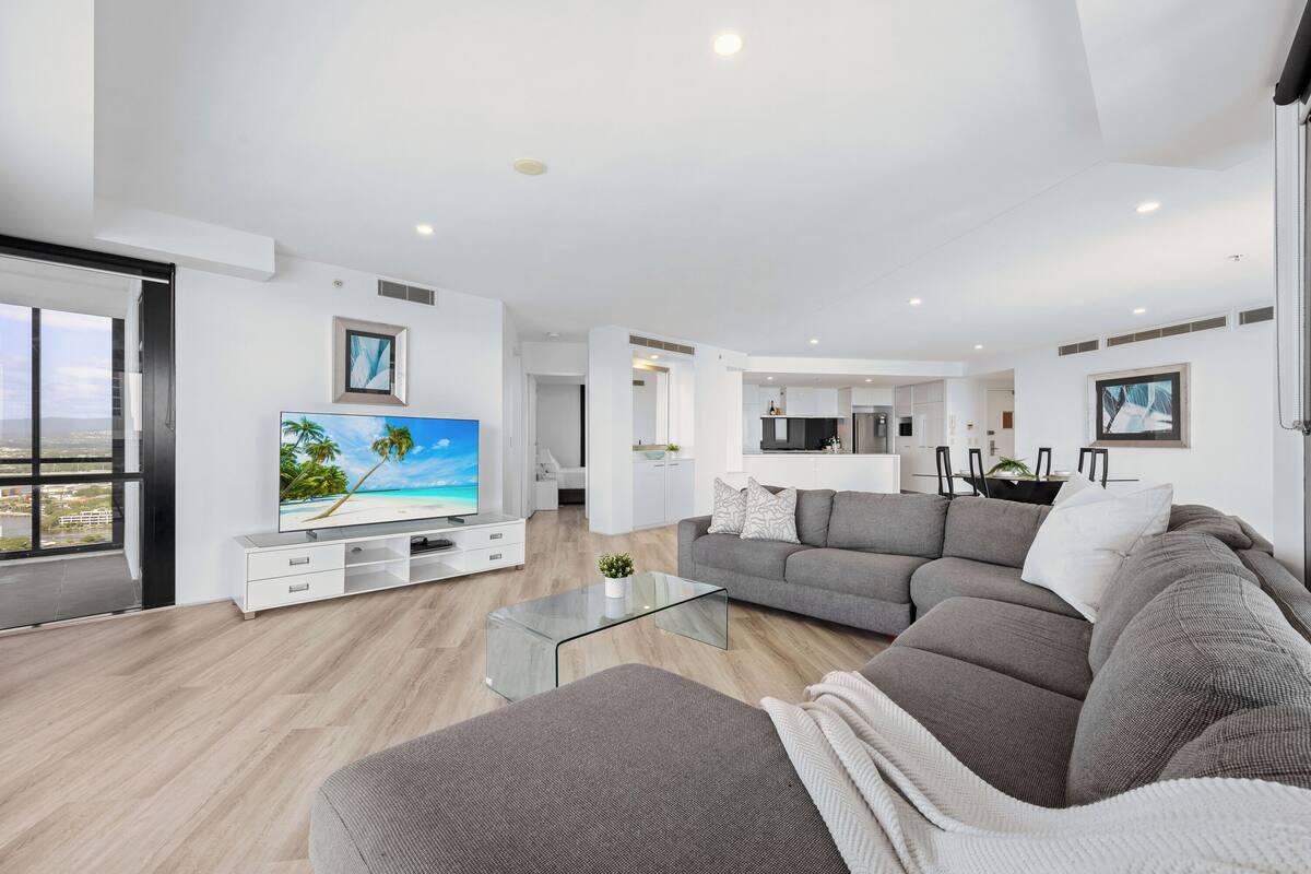 A modern living room with a large flat-screen TV, grey sofas, and a glass coffee table.