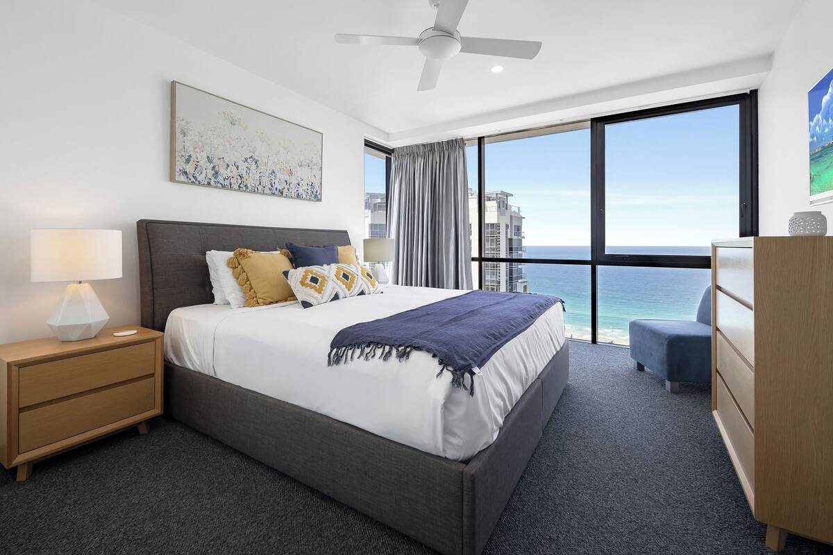 A modern hotel room with a large bed, a nightstand, a dresser, and a view of the ocean.