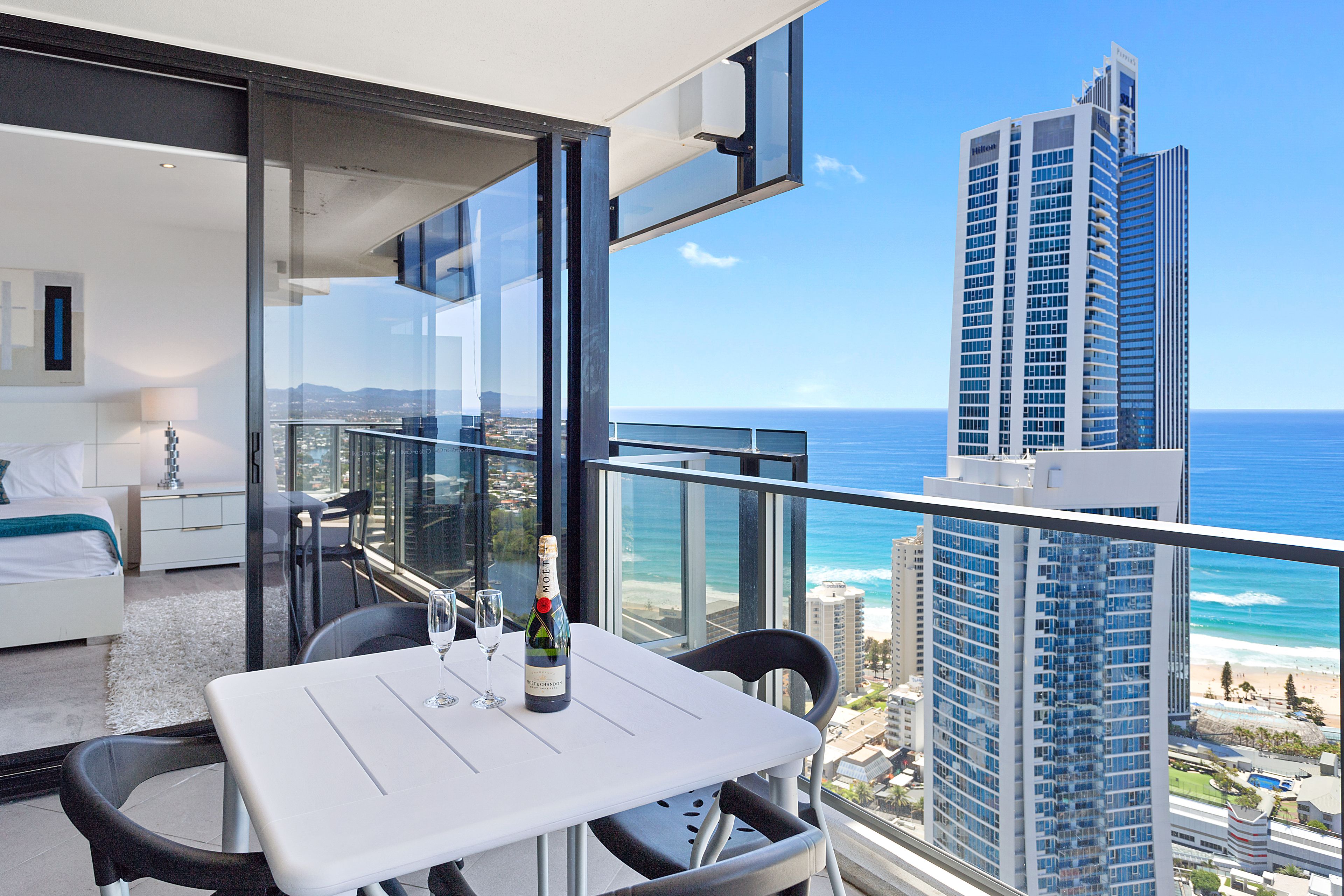 Un balcon avec une table et des chaises, un lit, et une vue sur l’océan et un grand immeuble.