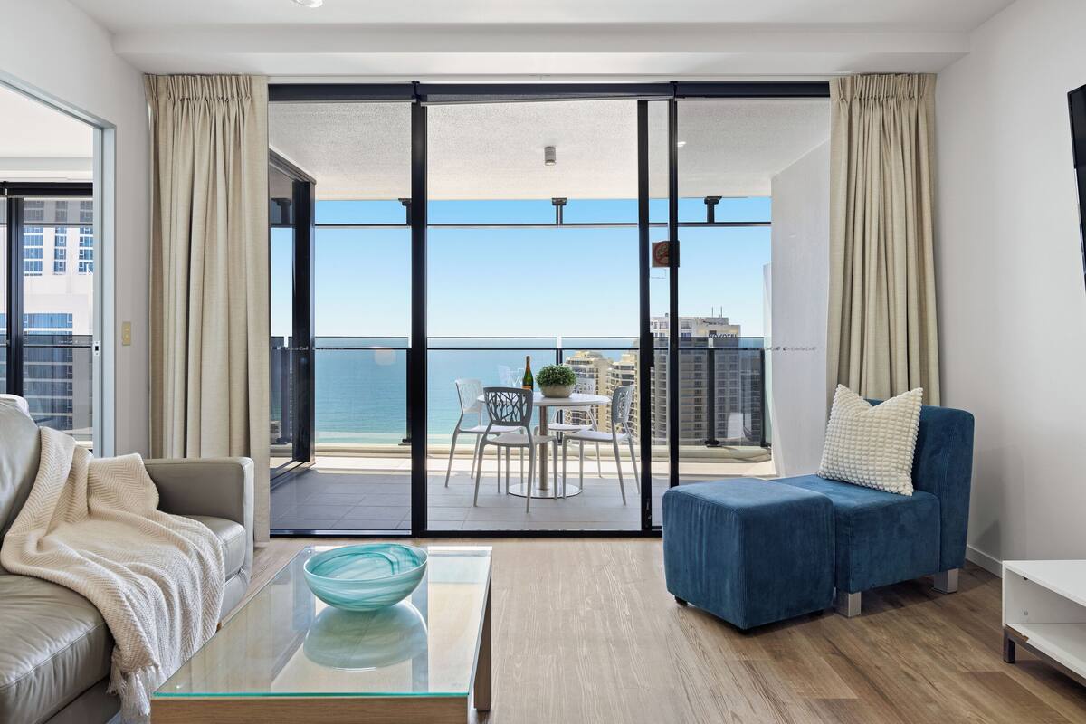 A modern living room with a glass coffee table, a blue sofa, and a view of the sea through large sliding doors.