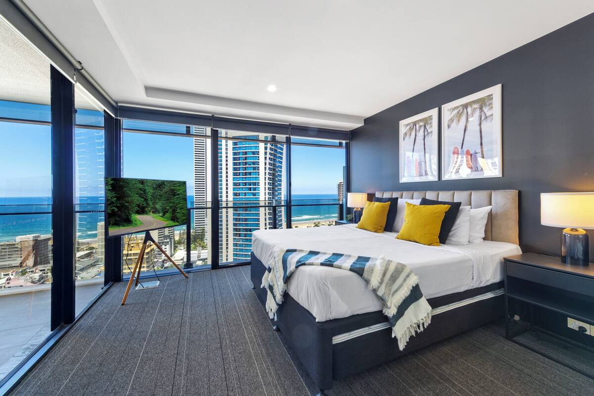 A modern bedroom with a large bed, a nightstand, and a view of the ocean through floor-to-ceiling windows.