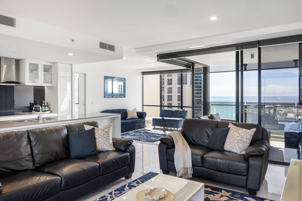 A modern living room with two black leather sofas, a coffee table, and a large window offering a city view.