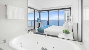 Ett modernt hotellrum med ett stort badkar, en säng med ett blått överkast och utsikt över havet genom stora fönster.