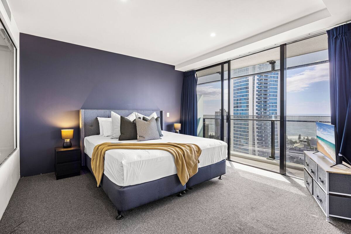 A modern hotel room with a large bed, a nightstand, a dresser, and a view of the city through large windows.
