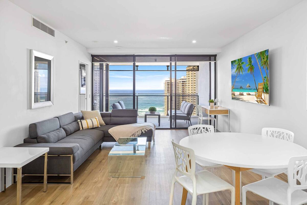 A modern living room with a large window, a gray sofa, a white dining table, and a painting of a beach scene.