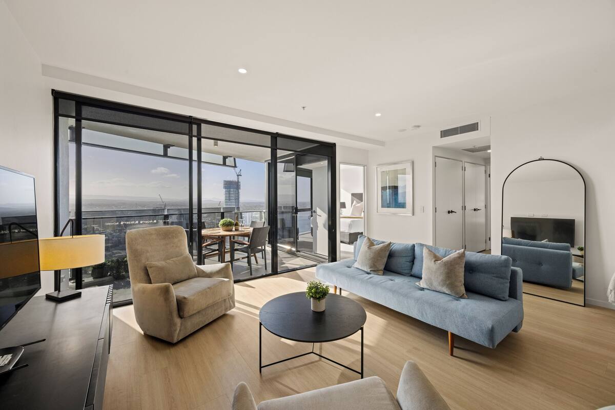 A modern living room with a sofa, armchair, and coffee table. There is a large window offering a view of a cityscape.