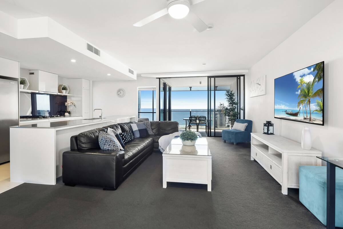 A modern living room with a black leather sofa, white coffee table, and a large window overlooking the ocean.