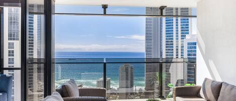 A balcony with wicker furniture, a potted plant, and a view of the ocean and city skyline.